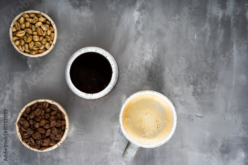 four ceramic cups with different stages of coffee: green and roasted beans and ready drink. On gray background