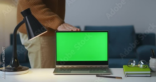 man is closing laptop with green screen in room at evening and turning off lamp, finished working, view on working table