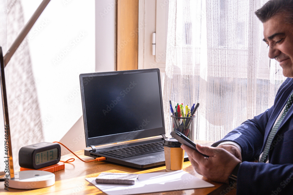 Fototapeta premium businessman at his desk with laptop and coffee working from home and smiling at what is sent to him on his mobile phone