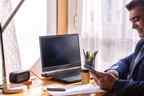 businessman at his desk with laptop and coffee working from home and smiling at what is sent to him on his mobile phone
