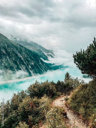 View through the fir trees to the clear turquoise mountain lake Schlegays covered with clouds and fog. Zillertal Alps, Mayrhofen, Austria. Vertical shot