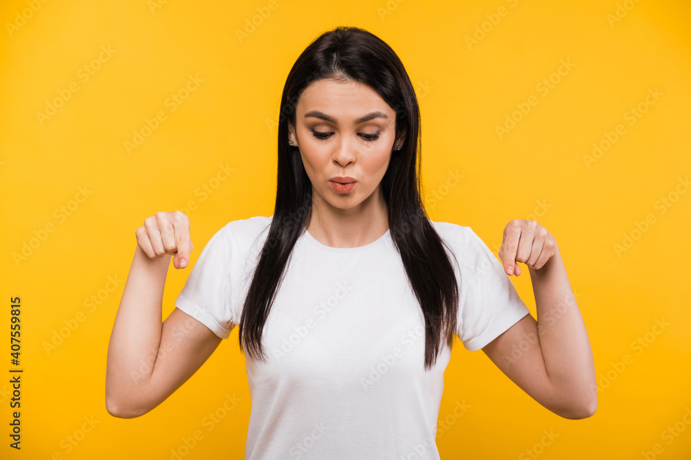 Pretty caucasian brunette girl in a white t-shirt, points fingers down with surprised look, workspace, isolated orange background Pretty caucasian brunette girl in a white t-shirt, points fingers down with surprised look, workspace, isolated orange background