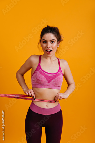 White young woman measuring her waiste. Lovely girl in sportswear holding measure tape. © Look!