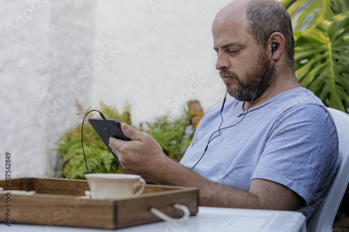 In an outdoor garden a man is looking intently at a digital tablet with headphones for listening.