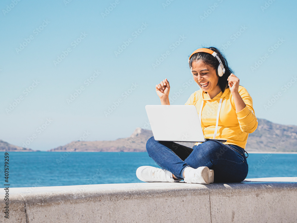 Foto Mujer latina joven feliz escuchando música en computadora portátil junto al mar de ropa amarrilla y audífonos 