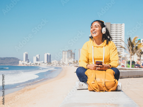 foto mujer joven sonriente escuchando música con audífonos junto al mar vestida de camisa amarilla  