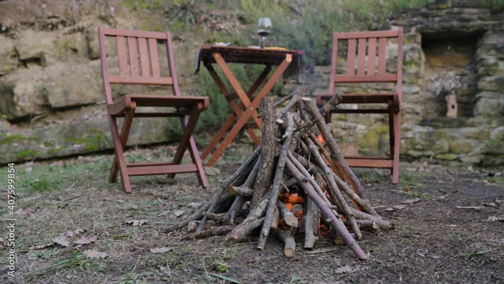 Close-up turning fire with pile of wood. Two wooden chairs and a table on background. Camping, outdoors concept. Close-up turning fire with pile of wood. Two wooden chairs and a table on background. Camping, outdoors concept.