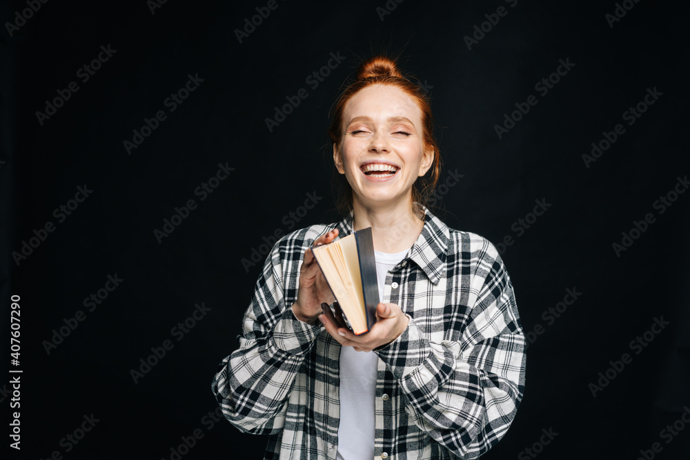 Laughing young woman college student standing holding opened book on isolated black background. Pretty redhead lady model emotionally showing facial expressions in studio