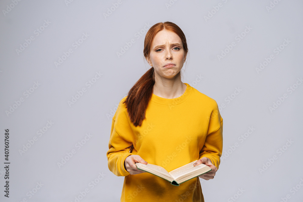 Obraz premium Confused displeased young woman college student holding book and crying on gray isolated background. Pretty redhead lady model wearing yellow sweater emotionally showing facial expressions.