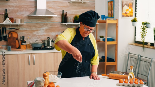 Elderly woman adding flour on table by hand sitting in the home kitchen. Retired senior baker with bonete and uniform sprinkling, sifting, spreading rew ingredients baking homemade pizza and bread. photo