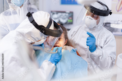 Orthodontist and assistant using drill on little girl cavity dressed in protective suit in the course of coronavirus. Stomatology team wearing ppe suit during covid19 doing procedure on child teeth.