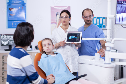 Pediatric dentist with tooth little girl digital radiography on tablet pc. Stomatologist explaining teeth diagnosis to mother of child in health clinic holding x-ray.