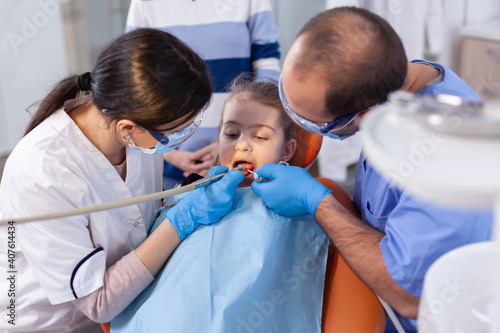 Dentist and assistant using instruments during dental treatment for little girl sitting on chair wearing big. Mother with her kid in stomatology clinic for teeth examine using modern instruments.