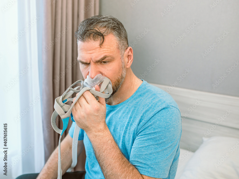 Unhappy shocked man with chronic breathing issues surprised by using CPAP machine sitting on the bed in bedroom. Healthcare, CPAP, Obstructive sleep apnea therapy, snoring concept Unhappy shocked man with chronic breathing issues surprised by using CPAP machine sitting on the bed in bedroom. Healthcare, CPAP, Obstructive sleep apnea therapy, snoring concept