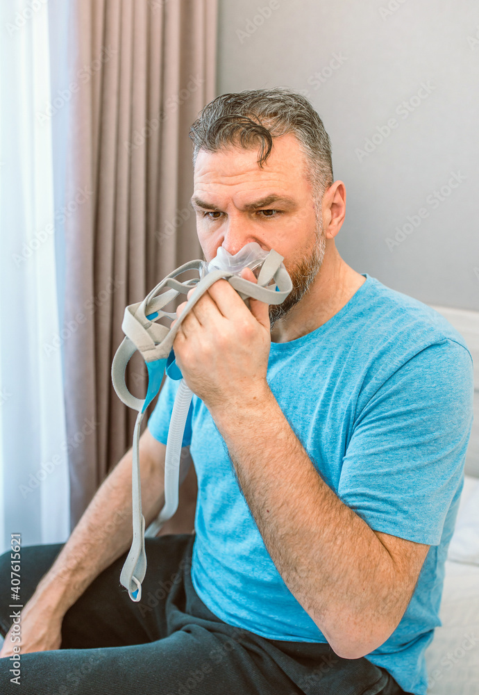 Unhappy shocked man with chronic breathing issues surprised by using CPAP machine sitting on the bed in bedroom. Healthcare, CPAP, Obstructive sleep apnea therapy, snoring concept Unhappy shocked man with chronic breathing issues surprised by using CPAP machine sitting on the bed in bedroom. Healthcare, CPAP, Obstructive sleep apnea therapy, snoring concept