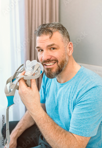 Mature happy man with chronic breathing issues considers using sitting on the bed in bedroom with CPAP machine. Healthcare, CPAP, snoring concept
