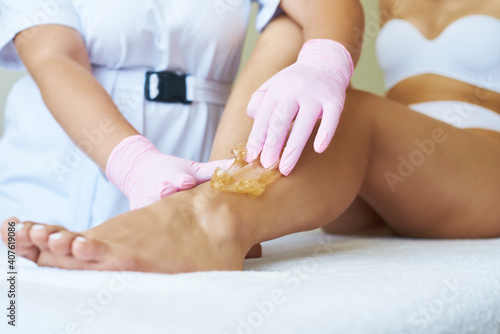 Lovely young beautician applies sugar paste to the client's leg. Hair removal with shugaring. Beautician depilating young woman