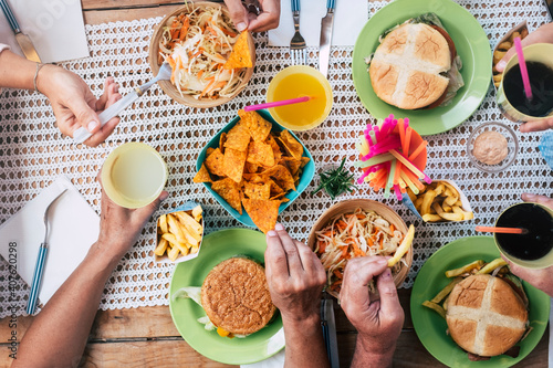 Close up of caucasian friends hands taking fast food hamburger and frech fries from the table - group of unrecognizable people eating and having fun together