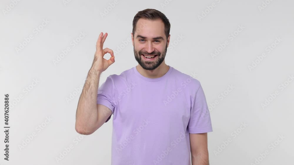 Smiling funny bearded young man 20s years old in pastel violet casual t-shirt isolated on white color background studio. People sincere emotions lifestyle concept. Showing ok okay gesture blinking Smiling funny bearded young man 20s years old in pastel violet casual t-shirt isolated on white color background studio. People sincere emotions lifestyle concept. Showing ok okay gesture blinking