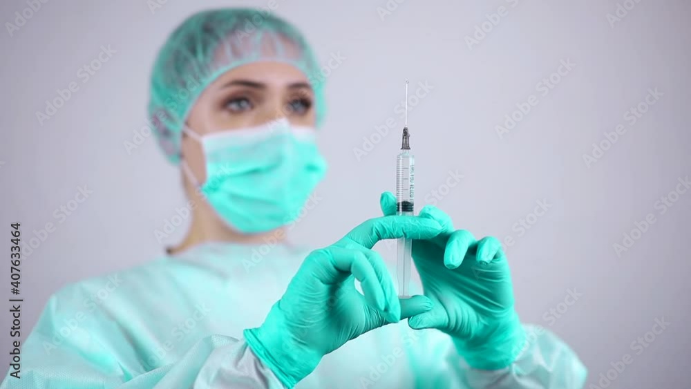 A nurse wearing a medical mask and protective gloves looks at the needle of a syringe for injection A nurse wearing a medical mask and protective gloves looks at the needle of a syringe for injection