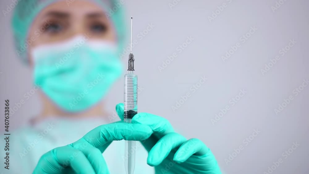 A nurse wearing a medical mask and protective gloves looks at the needle of an injection syringe dripping with a drop A nurse wearing a medical mask and protective gloves looks at the needle of an injection syringe dripping with a drop