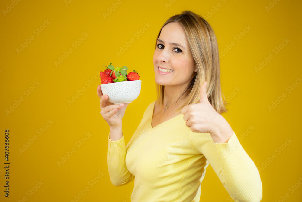 Beautiful cheerful young woman standing and holding glass bowl with strawberries with thumb up over yellow background Beautiful cheerful young woman standing and holding glass bowl with strawberries with thumb up over yellow background