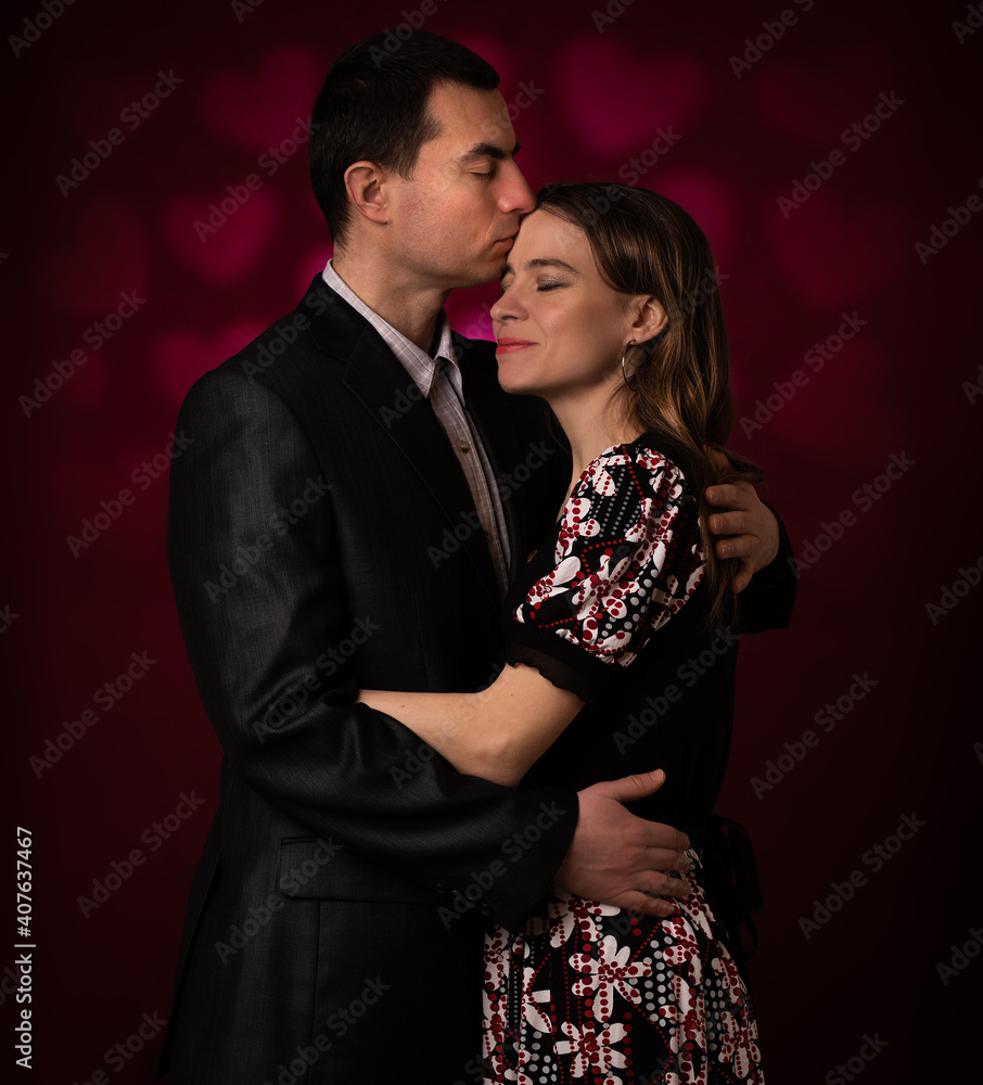 Young man and woman embracing, kissing and smiling against red wall with heart shapes