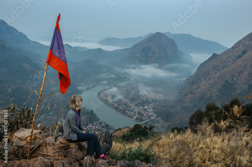 Chill out after morning hike to the peak of the mountain.Female traveler sitting on a rock in Laos, Lao national flag.Sunrise watching.Amazing view of Nam Ou river,mountains. Relaxation positive vibes