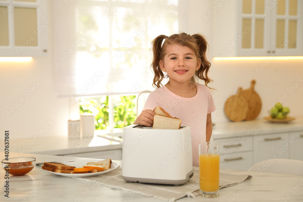 Cute little girl putting slice of bread into toaster at table in kitchen