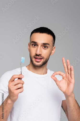 smiling hispanic man holding toothbrush while showing ok gesture isolated on grey