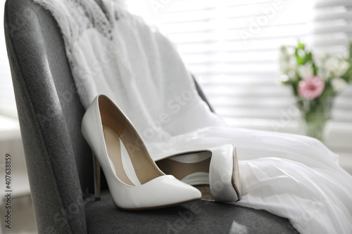 Pair of white high heel shoes and wedding dress on chair indoors © New Africa