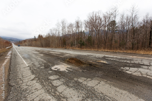 Bad Russian asphalt road. The asphalt road is full of holes and cracks among dried autumn trees.