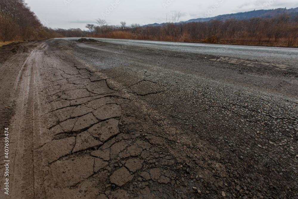 Bad Russian asphalt road. The asphalt road is full of holes and cracks among dried autumn trees.