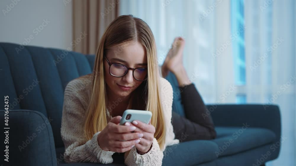 Young attractive woman with glasses texting a message on the smartphone while chatting at home on couch. Smiling. Online in social media. Close up. Slow motion Young attractive woman with glasses texting a message on the smartphone while chatting at home on couch. Smiling. Online in social media. Close up. Slow motion