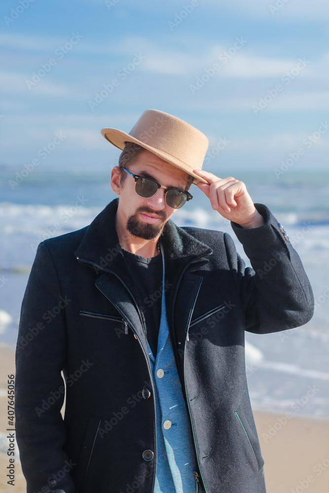 Young black beard man in blue jacket and black coat and black sunglasses touching brown hat looking at camera enjoying sunlight at the seaside on sunny day. Male fashion, hipster guy, male emotions.