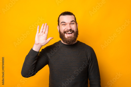 Photo of joyful happy bearded hipster man making hello gesture over yellow background.
