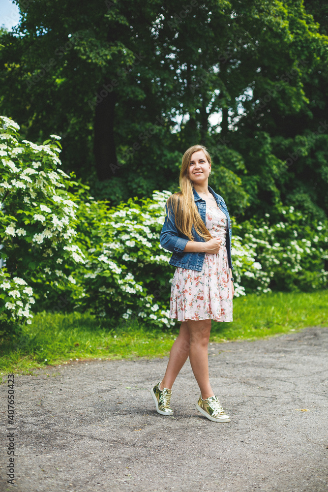 A young woman of European appearance with long blond hair, dressed in a short dress, stands against the background of white flowering bushes. Sunny spring day. Natural female beauty A young woman of European appearance with long blond hair, dressed in a short dress, stands against the background of white flowering bushes. Sunny spring day. Natural female beauty