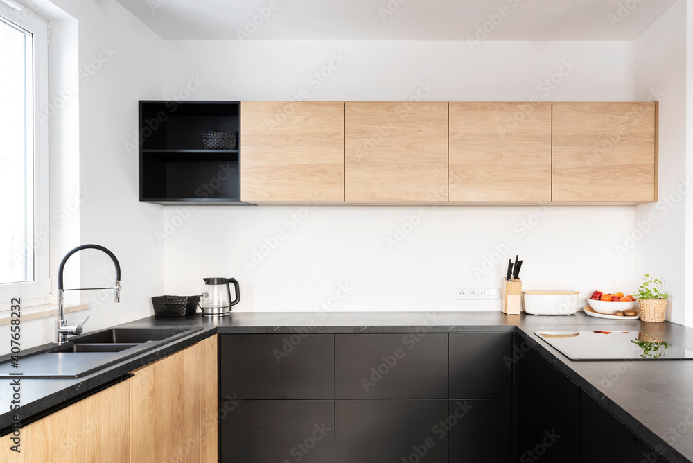 Minimalism in modern kitchen with blacj countertop and induction hob and wooden furniture. Interior of kitchen with window and stylish sink and faucet. Minimalism in modern kitchen with blacj countertop and induction hob and wooden furniture. Interior of kitchen with window and stylish sink and faucet.