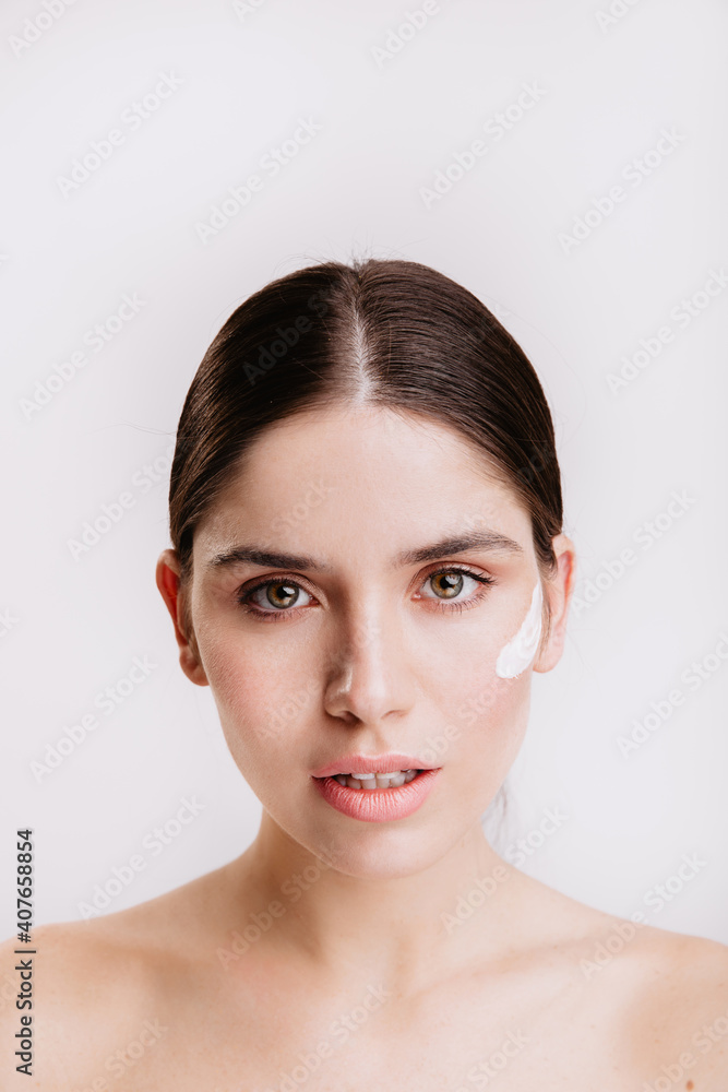 Close-up portrait of green-eyed dark-haired woman with healthy skin and cream on her face. Girl without makeup looking at camera on white background Close-up portrait of green-eyed dark-haired woman with healthy skin and cream on her face. Girl without makeup looking at camera on white background
