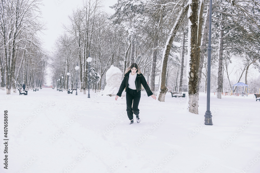 A beautiful young girl in warm clothes walks, plays and poses against the background of a winter snow-covered park and a playground