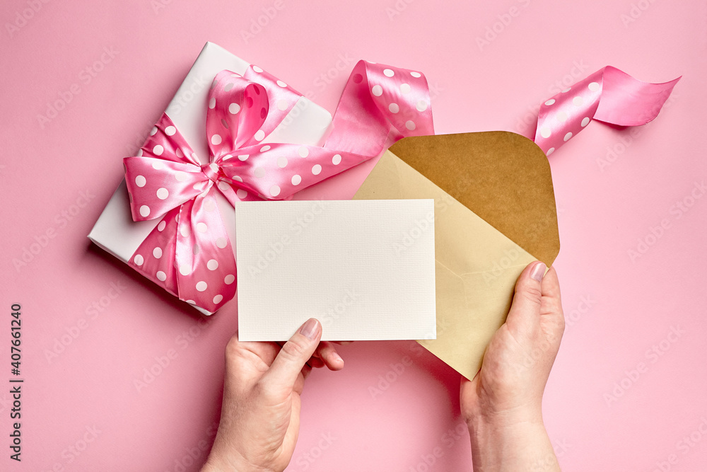 Greeting card mockup with gift box with bow in female hands on pink paper background Greeting card mockup with gift box with bow in female hands on pink paper background