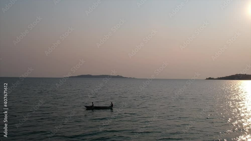 Sunset on deep blue waters of the lake with two men on a traditional boat, Makoro, paddling from right to left, pink sunset and the small mountain on the distant horizon. Lake Tanganyika. Sunset on deep blue waters of the lake with two men on a traditional boat, Makoro, paddling from right to left, pink sunset and the small mountain on the distant horizon. Lake Tanganyika.
