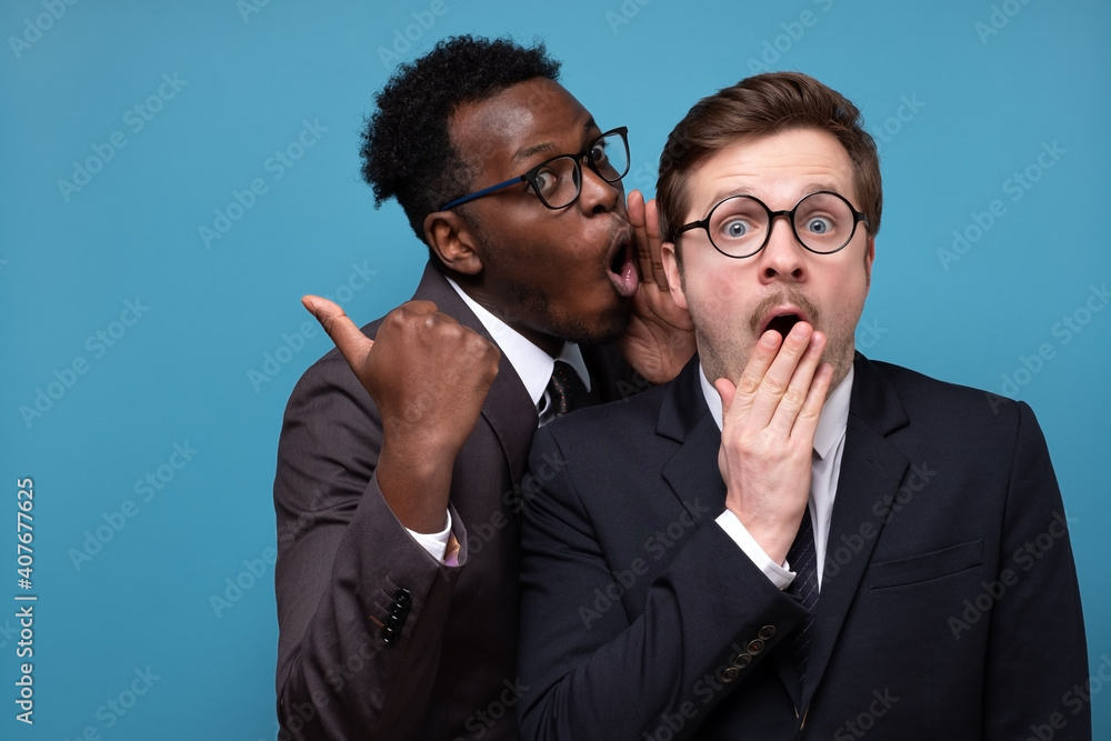 African man whisperingto his caucasian coworker ear telling him something secret. Studio shot on blue wall. Human emotions facial expressions African man whisperingto his caucasian coworker ear telling him something secret. Studio shot on blue wall. Human emotions facial expressions
