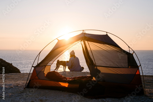 Woman and her pet dog sit at beach next to tent, solitude camping in nature. Sunset shot of young woman have self exploration time and connection to nature. Tranquility and mindfulness