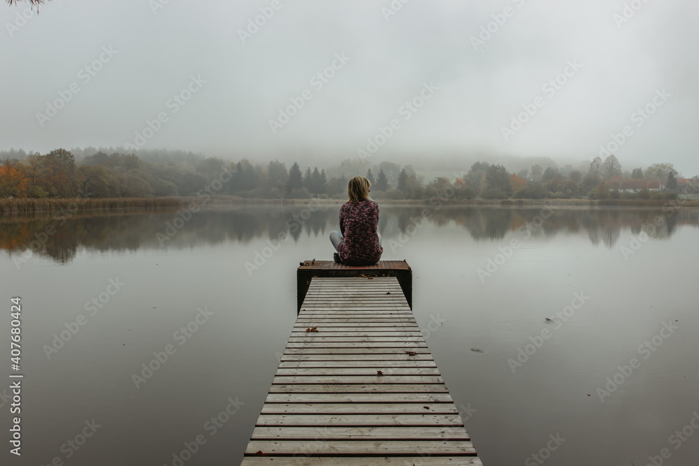 Calm morning relaxation by the foggy pond. Sitting woman in the countryside. Intended female mysterious atmosphere. Autumn afternoon in the nature. Silence. Wooden pier on a pond.