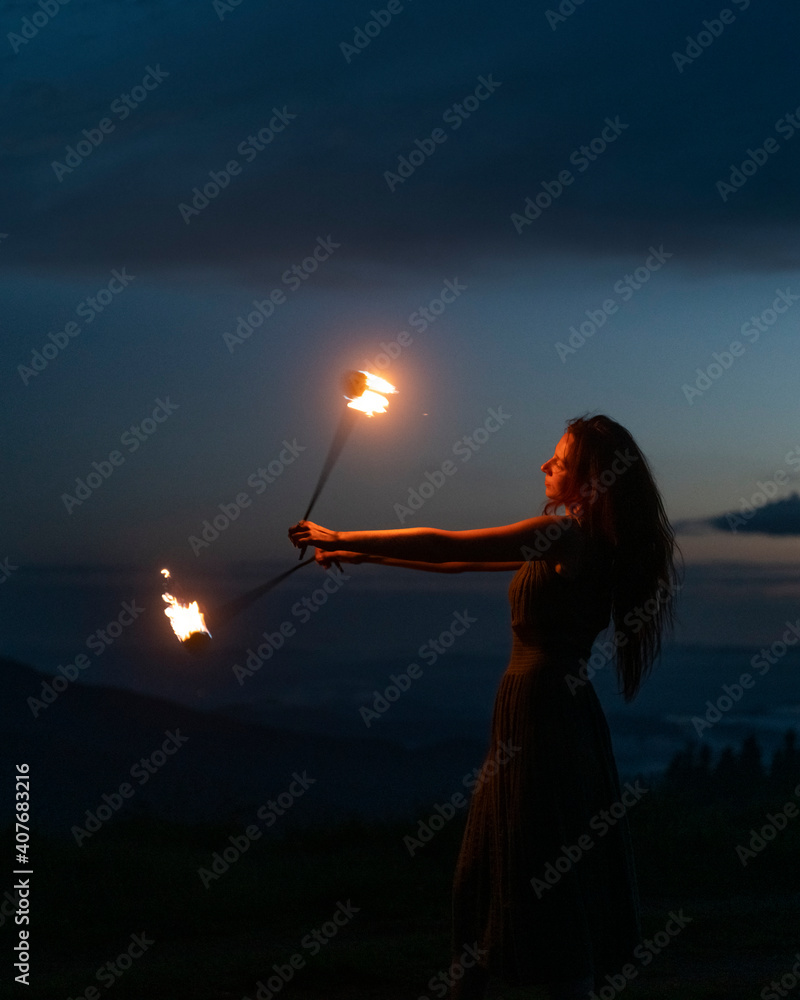 Graceful elegant red haired female fireshow performer making fire dance with two metal fans lit with flames on night mountains meadow