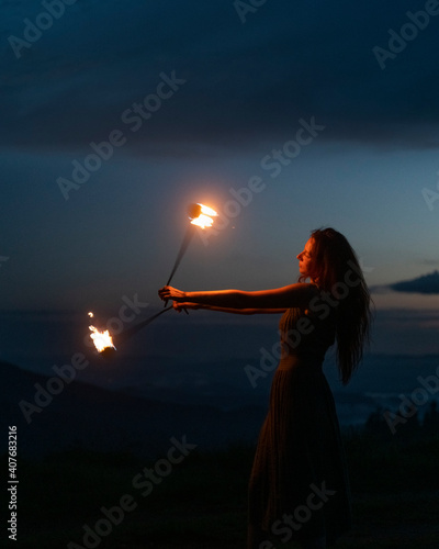 Graceful elegant red haired female fireshow performer making fire dance with two metal fans lit with flames on night mountains meadow photo