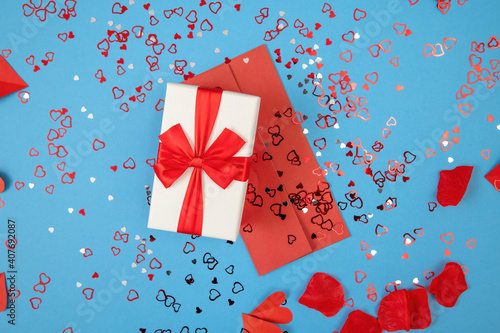 Presnet with envelope lying on blue background with glitters from directly above. Box with ribbon and red postcard on colorful desk in flat lay. Gift and card with heart sequins. photo