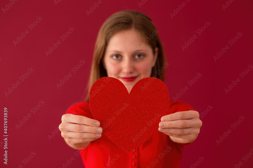 Blurred female showing heart to the camera on red background. Blonde girl holding glittered symbol of love on colorful backdrop. Smiling woman on valentine's day from close up. Blurred female showing heart to the camera on red background. Blonde girl holding glittered symbol of love on colorful backdrop. Smiling woman on valentine's day from close up.