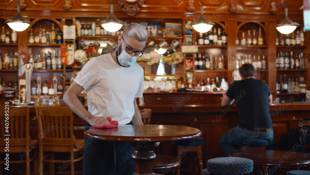 Waiter wearing protection face mask in apron cleaning table with disinfectant spray Waiter wearing protection face mask in apron cleaning table with disinfectant spray